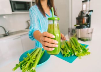 Woman in kitchen offering bottle of freshly pressed juice in juicer machine.