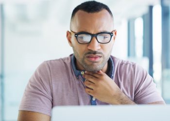 Man in front of lap top at office with uncomfortable look on his face as he tries to clear his throat; he is touching his throat with one hand