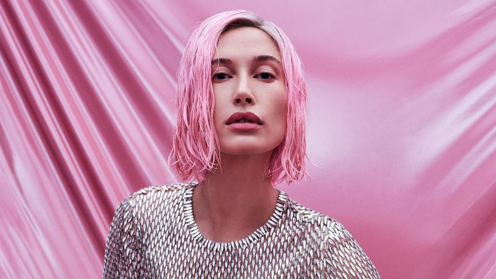 Hailey Bieber photographed for Allure Magazine by Zoey Grossman. She has a short, bubblegum pink bob and is wearing a silver bodysuit that looks like chainmail. She is sitting on the ground in front of a pink cloth backdrop with one leg propped up.