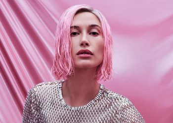 Hailey Bieber photographed for Allure Magazine by Zoey Grossman. She has a short, bubblegum pink bob and is wearing a silver bodysuit that looks like chainmail. She is sitting on the ground in front of a pink cloth backdrop with one leg propped up.