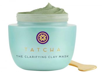 A small gold face mask spoon next to an open light blue jar filled with the green Tatcha The Clarifying Clay Mask on a white background