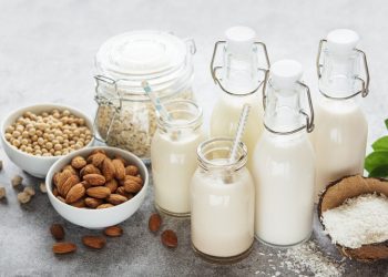A variety of plant-based milks in bottles against a gray background. Nuts, seeds, oats, coconut flakes in the shell, and green leaves also are shown.