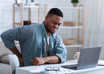 Man sitting at desk holding lower back and grimacing.