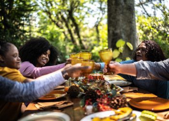 A family eating a meal near leafy green trees outdoors; three out of five people are seen close up during a toast