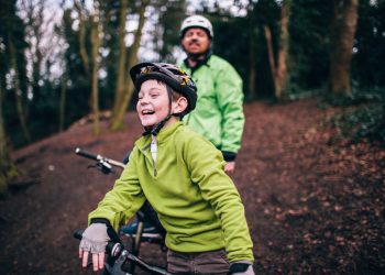 Father behind laughing son, both on bikes on a trail with trees behind them