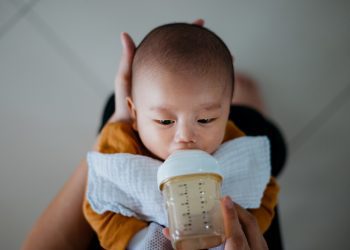 Parent cradling infant being fed formula with one hand behind head and other hand holding bottle