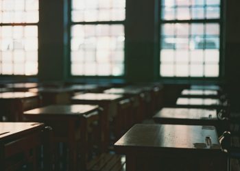 A classroom with several rows of empty desks and chairs in front of large-multipaned windows