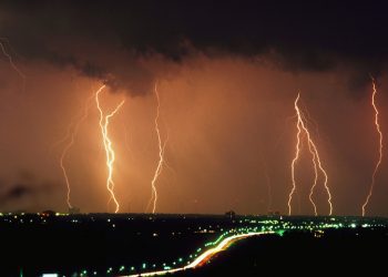 photo of a stormy night sky with multiple flashes of lightning spiking down from dark clouds over city lights and a blurry line of highway lights