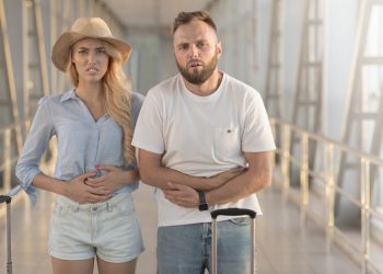 Couple standing in an airport terminal looking sick; bearded man has arms wrapped around his stomach and woman with long blond hair also has her hands on her stomach