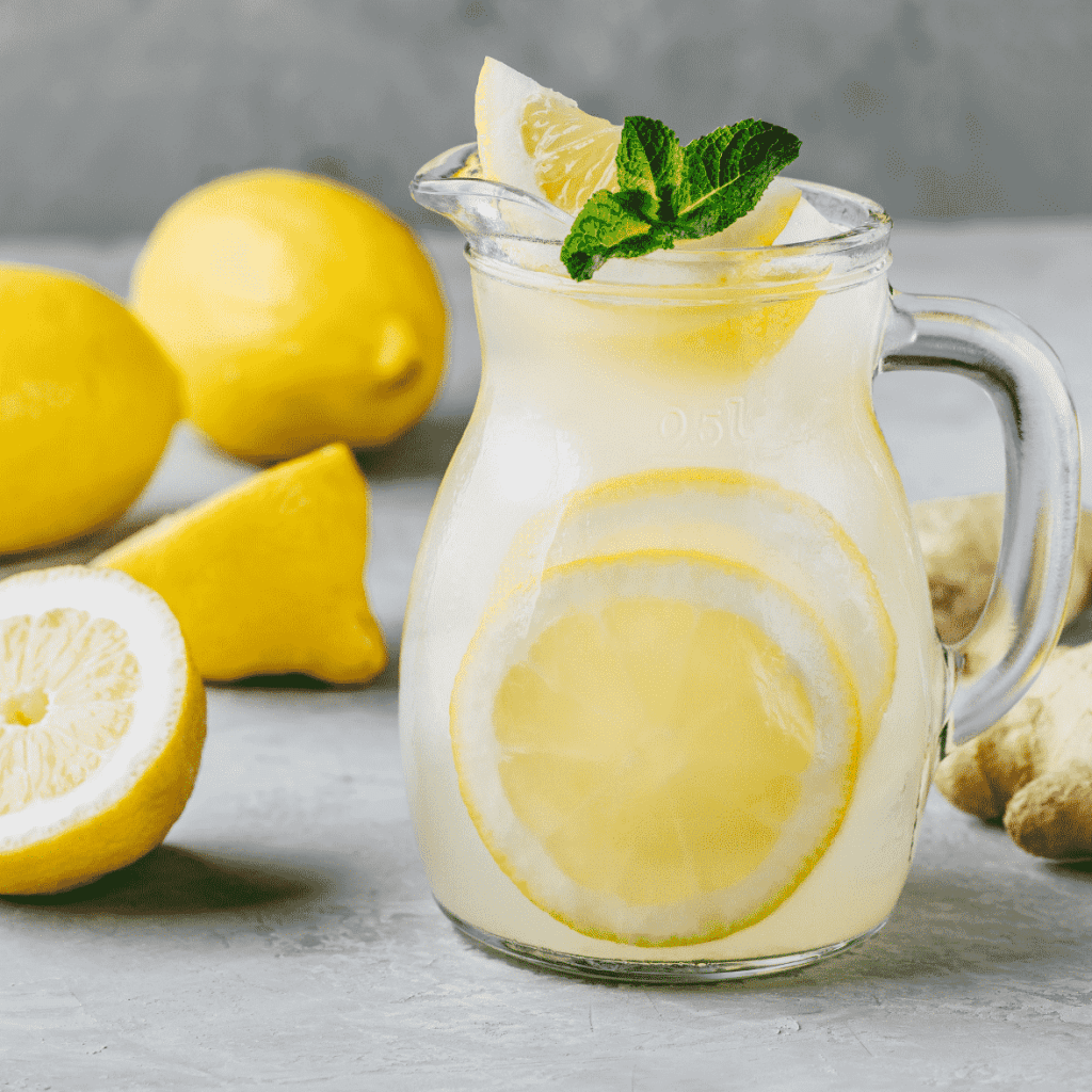 Glass pitcher of lemon water, garnished with a green leaf on top. Cut up yellow lemons in the background for style.