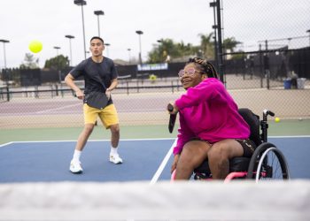 A man on a pickleball court wearing a dark tee shirt and yellow shorts and a woman in a wheelchair swinging back her racquet to hit the ball; she's wearing a bright pink top