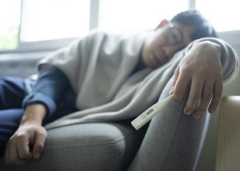 Man wearing glasses and holding thermometer in one hand lying on a couch with a blanket wrapped around him; concept is feeling sick