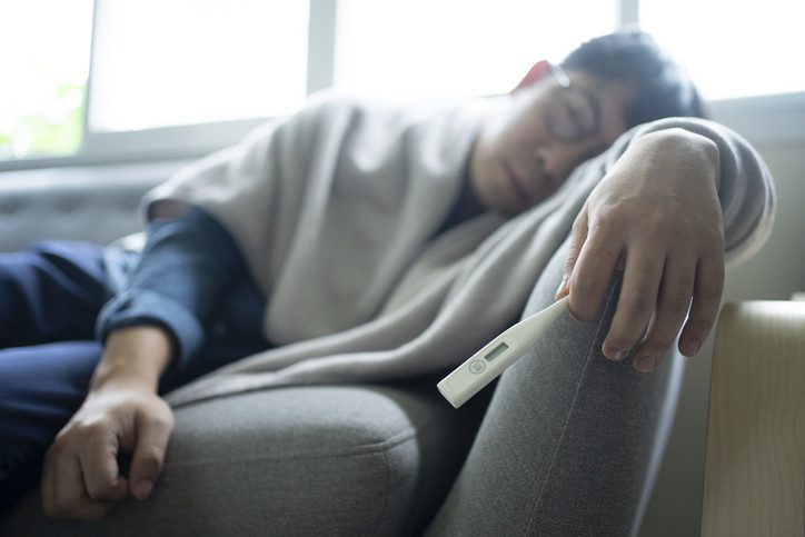 Man wearing glasses and holding thermometer in one hand lying on a couch with a blanket wrapped around him; concept is feeling sick