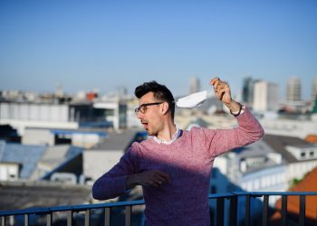 Man with dark hair and glasses standing outside near a railing with a cityscape in the background and ripping off his medical mask